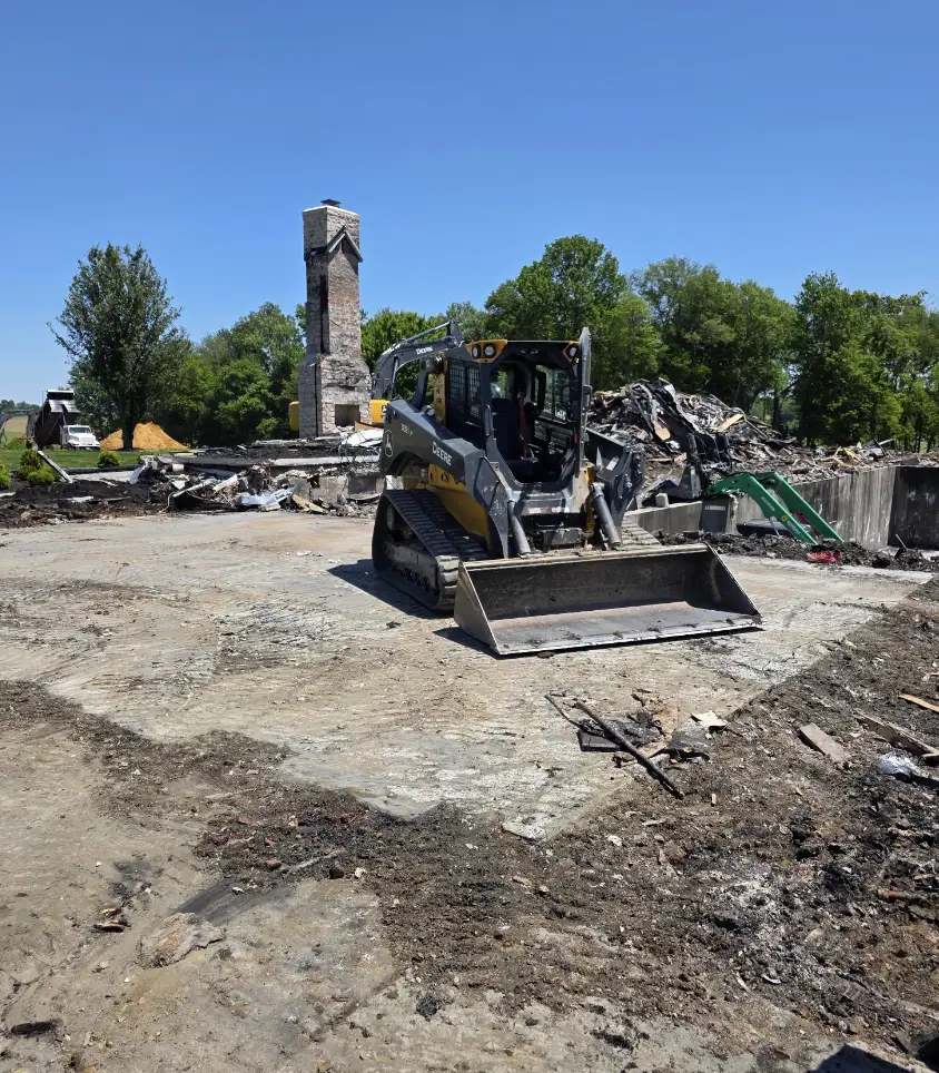 Tracked skid steer on a cleared slab after a house demolition, stone chimney behind
